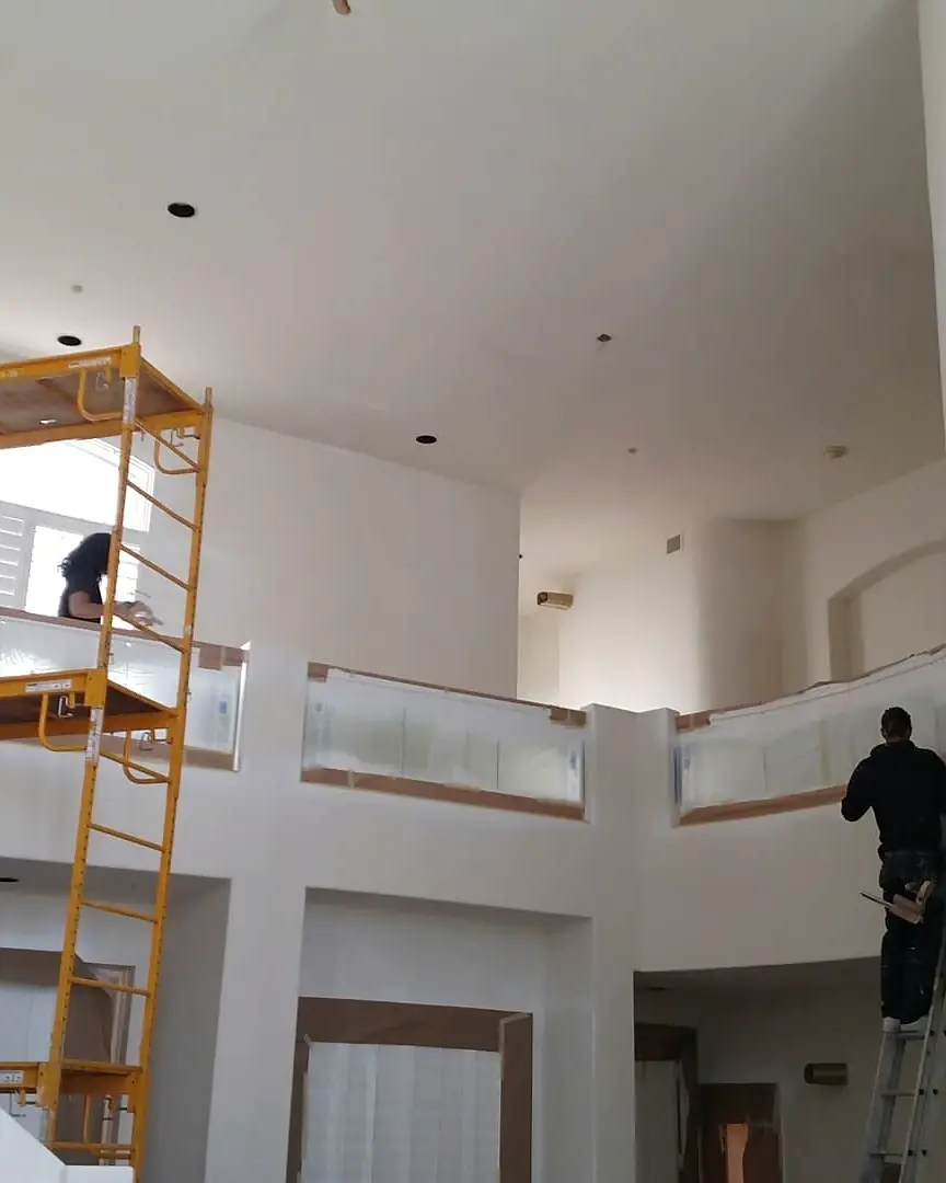 Painters working from a scaffold and ladder inside a tall white residential interior.
